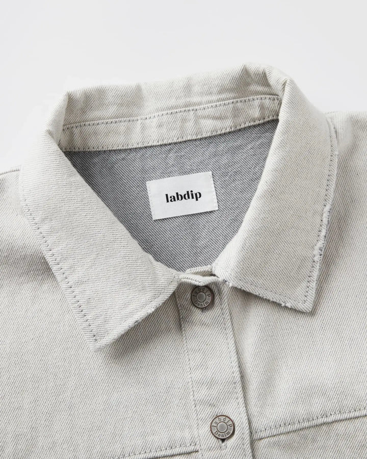 Col chemise en denim non teint couleur granit clair avec coutures apparentes et boutons métalliques, tissu 100% coton au toucher structuré et naturel.