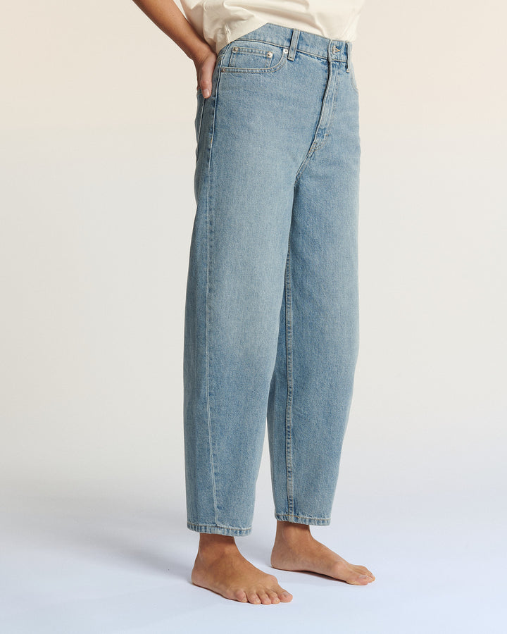 Jean en denim bleu clair pour femme, coupe droite et ample, adapté à toutes les morphologies, porté avec un t-shirt blanc, fond neutre beige.