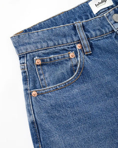 Détail d’un jean en denim bleu clair 100 % coton avec couture contrastante et poches renforcées par rivets en cuivre, coupe droite adaptée à toutes morphologies.