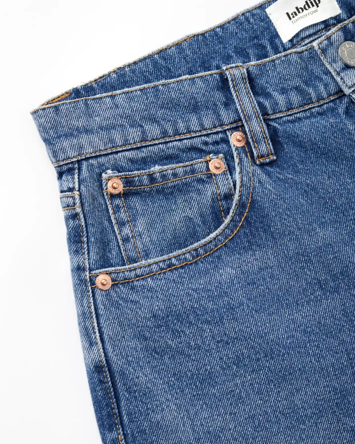 Détail d’un jean en denim bleu clair 100 % coton avec couture contrastante et poches renforcées par rivets en cuivre, coupe droite adaptée à toutes morphologies.