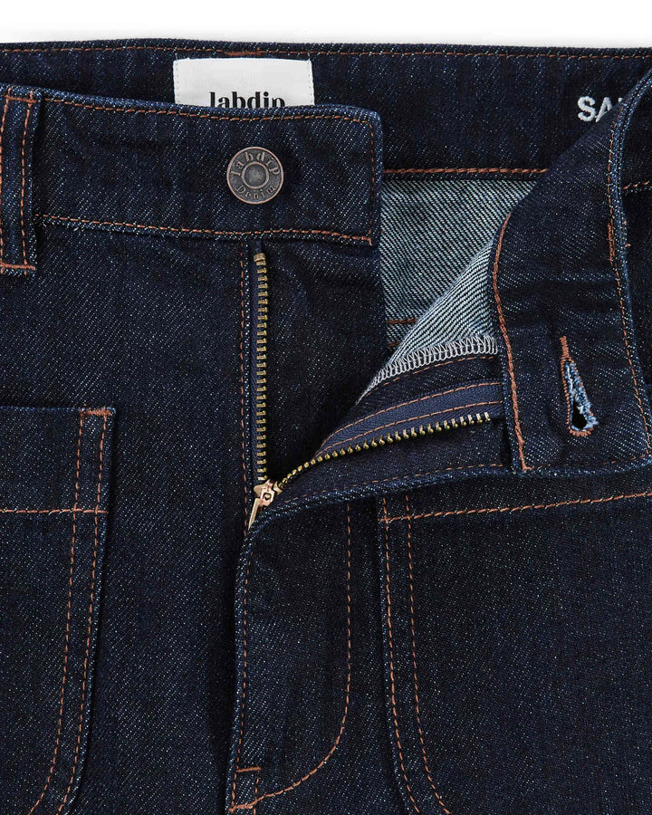 Jean femme en denim brut foncé 100% coton avec une coupe droite et un tissu épais et robuste, fermeture éclair métal et coutures apparentes couleur cuivrée.