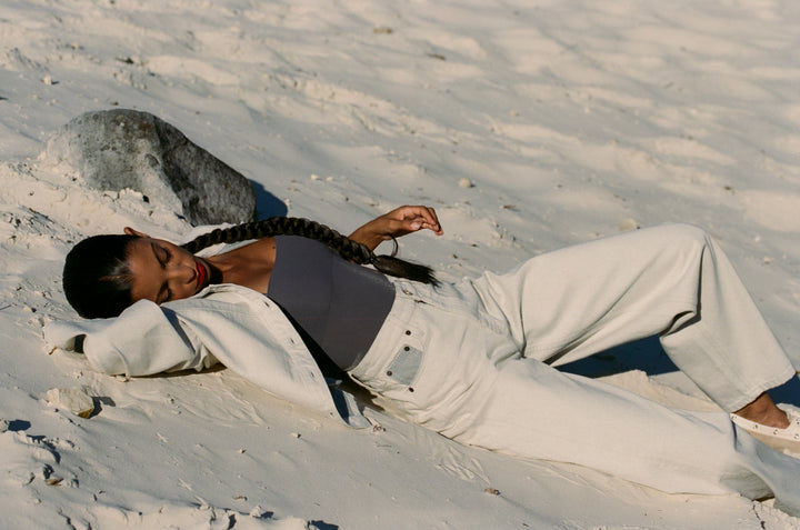 Femme allongée sur le sable clair portant un jean large blanc en coton avec un haut gris et une veste assortie, dans une ambiance détendue et naturelle.