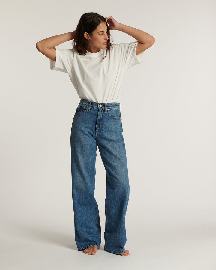 Femme portant un jean droit en denim bleu moyen avec un t-shirt blanc simple, posant pieds nus dans un studio lumineux au fond neutre.